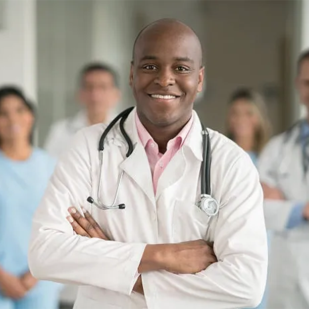 A middle aged bald black doctor wearing a white doctor's coat and a stethoscope is standing with his arms crossed in the foreground with his medical team blurred out in the background