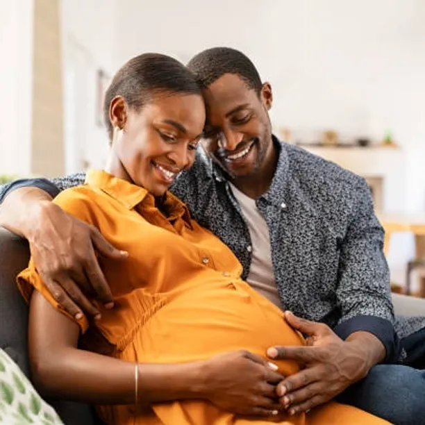 A smiling black couple is sitting. The woman is wearing a mustard yellow button down dress with collar and is looking down at her pregnant belly with her hands framing the outside.The male partner is sitting next to her with one hand wrapped around her shoulder and the other hand resting on her hands on her belly. He is wearing a blue patterned button up shirt over a white t-shirt and blue denim jeans.