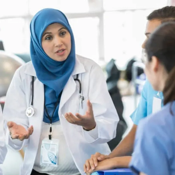 A doctor who is a woman wearing a blue hijab, white doctors coat, and stethoscope is shown in mid conversation with others